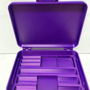 May include: A rectangular purple storage case with an open hinged lid. The interior features a molded base with various compartments, including three small horizontal slots, two square sections, and a long horizontal channel.