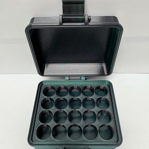 Puede incluir: Un estuche rectangular verde oscuro abierto, con tapa abisagrada y asa. El interior del estuche tiene 20 compartimentos circulares. El estuche está diseñado para guardar objetos pequeños.