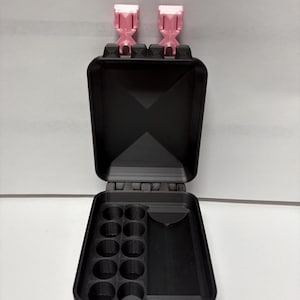 May include: A black rectangular storage case with a hinged lid and two light pink latches. The interior features a section with ten circular cylindrical compartments arranged in two rows and a separate rectangular open storage compartment.