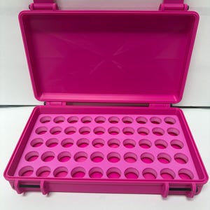 Può includere: Una custodia rettangolare rosa acceso con coperchio incernierato. L'interno presenta un inserto in schiuma con quarantotto fori circolari disposti a griglia, progettato per organizzare piccoli oggetti all'interno del contenitore rigido.