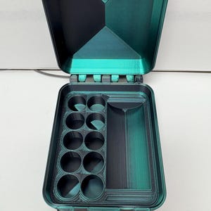 Könnte beinhalten: Ein dunkelgrüner und schwarzer 3D-gedruckter Aufbewahrungskoffer mit offenem Deckel. Das Innere verfügt über acht kreisförmige Fächer in zwei Spalten auf der linken Seite und einen einzelnen rechteckigen Schlitz auf der rechten Seite.