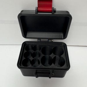 May include: A black rectangular storage case with an open lid and a red latch. The interior features a custom insert with multiple circular cutouts designed for holding small cylindrical items. The exterior has a textured finish and a hinged design.