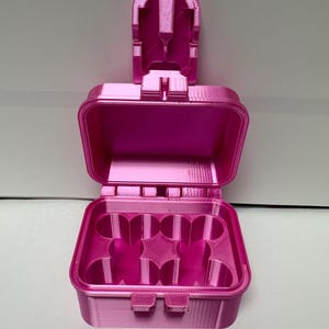 Puede incluir: Una caja de almacenamiento rectangular de color rosa brillante impresa en 3D con la tapa abierta. La base interior presenta cuatro compartimentos circulares dispuestos en cuadrado y la tapa incluye un mecanismo de cierre vertical.