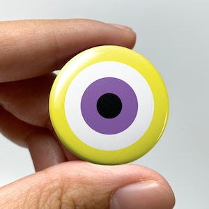 May include: A round button with a yellow background and a stylized eye design. The eye features concentric circles in white, purple, and a black pupil. The button is held between fingers.