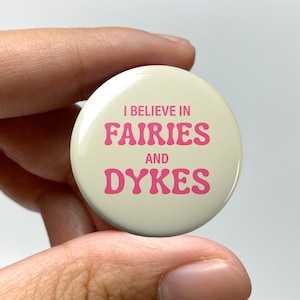 May include: A white button with pink text that reads "I believe in fairies and dykes".