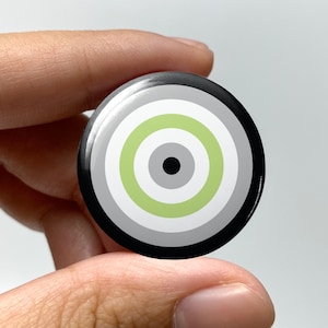 May include: A black and white button with a green, gray, and black concentric circle design. The button is a circular shape and has a glossy finish.