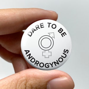 May include: White button with black text that reads "Dare to be Androgynous". The button features a stylized image of a circle with a cross inside, representing the intersection of male and female symbols.