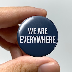 May include: A navy blue button with the white text "WE ARE EVERYWHERE." The button is round and has a glossy finish. It is held between fingers against a light background.