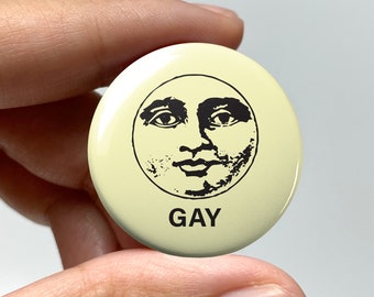以下が含まれることがあります： 男性の顔の白黒画像が描かれた黄色のボタンで、下に「GAY」という文字が印刷されています。