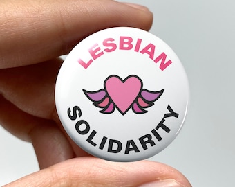 May include: A white button with pink and purple wings and a pink heart in the center. The text "Lesbian Solidarity" is written in a black circle around the heart.