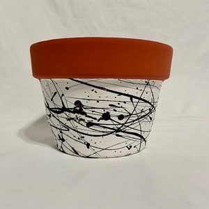 May include: A terracotta planter with a white base and black paint splatters and lines. The planter is a simple, round shape.