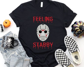 May include: Black t-shirt with a white hockey mask graphic and the text "Feeling Stabby" in red dripping letters. The shirt is laid flat on a white surface with a pair of black and white checkered Vans shoes, a pair of blue jeans, and a black mug with a spider web design.