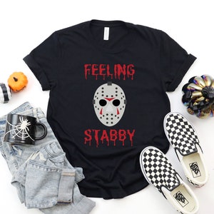 May include: Black t-shirt with a white hockey mask graphic and the text "Feeling Stabby" in red dripping letters. The shirt is laid flat on a white surface with a pair of black and white checkered Vans shoes, a pair of blue jeans, and a black mug with a spider web design.