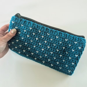 May include: A rectangular zipped pouch made of teal crochet yarn featuring a repeating pattern of small white V-shaped stitches. The pouch has a black zip closure along the top edge and a textured, hand-stitched appearance throughout.