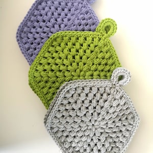 May include: Three hexagonal crochet potholders in lavender, green, and grey. Each has a loop for hanging. The textured stitch creates a puffy effect. These handmade kitchen accessories add charm. The potholders are ideal for protecting surfaces from heat.