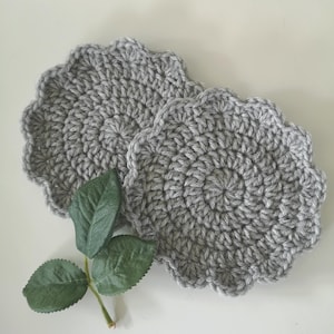 May include: Two grey, crocheted coasters with a scalloped edge. The coasters are circular and have a textured appearance. A sprig of green leaves rests near the coasters, adding a touch of natural colour.