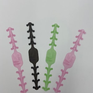 May include: Four ear savers in different colors: pink, black, green, and light green. The ear savers are made of plastic and have a textured surface. They are designed to help keep face masks from pulling on the ears.