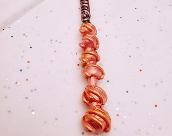 May include: A purple and gold wand with a pink and gold swirl design. The wand is on a white background with glitter.
