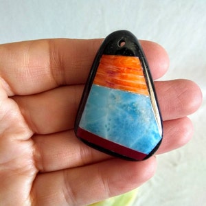 May include: A triangular pendant with a black top, orange and blue center, and red bottom. The pendant has a hole at the top for a necklace. The pendant is held in a hand.