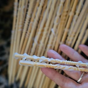May include: Two wooden wands with white glue applied to them. The wands are held in a hand with a ring on the finger. The background is a blurred image of a bunch of other wooden wands.