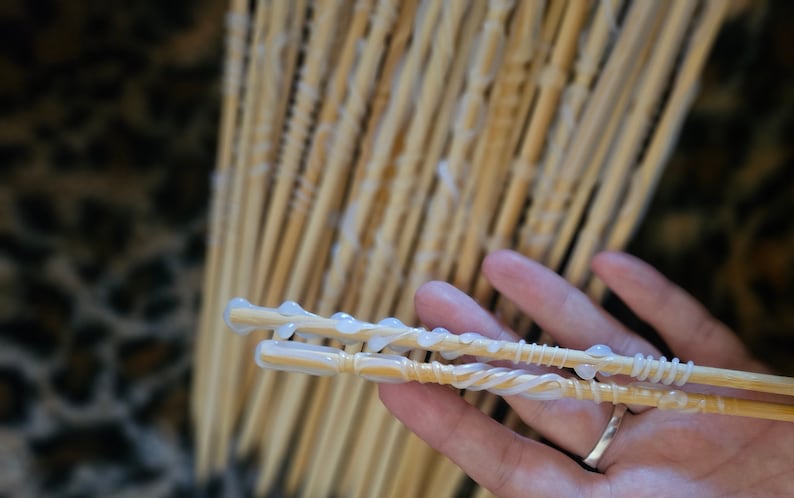 May include: Two wooden wands with white glue applied to them. The wands are held in a hand with a ring on the finger. The background is a blurred image of a bunch of other wooden wands.