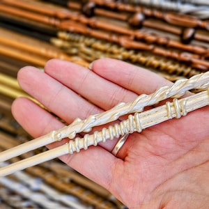 May include: Two silver wands with intricate designs. The wand on top has a twisted design, while the wand on the bottom has a series of rings. Both wands are held in a hand.