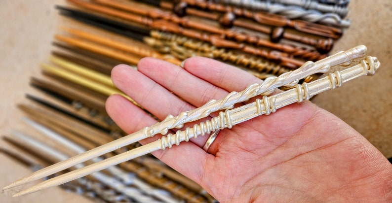 May include: Two silver wands with intricate designs. The wand on top has a twisted design, while the wand on the bottom has a series of rings. Both wands are held in a hand.