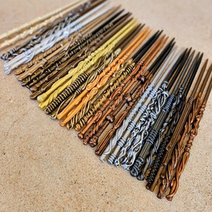 May include: A collection of 25 wooden wands in various colors, including gold, silver, brown, and black. The wands are arranged in a row, with the tips pointing to the right. The wands are all different lengths and have different designs.
