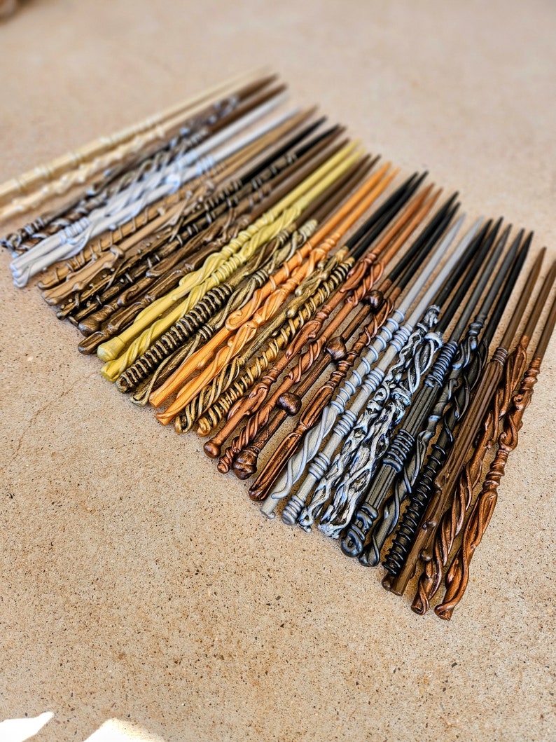 May include: A collection of 25 wooden wands in various colors, including gold, silver, brown, and black. The wands are arranged in a row, with the tips pointing to the right. The wands are all different lengths and have different designs.