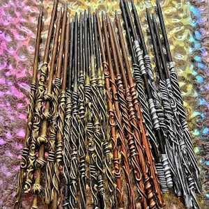 May include: A collection of 20 intricately designed wands, each with a unique pattern and color. The wands are made of a dark brown, copper, silver, and gold material. They are arranged in a fan shape on a textured surface.