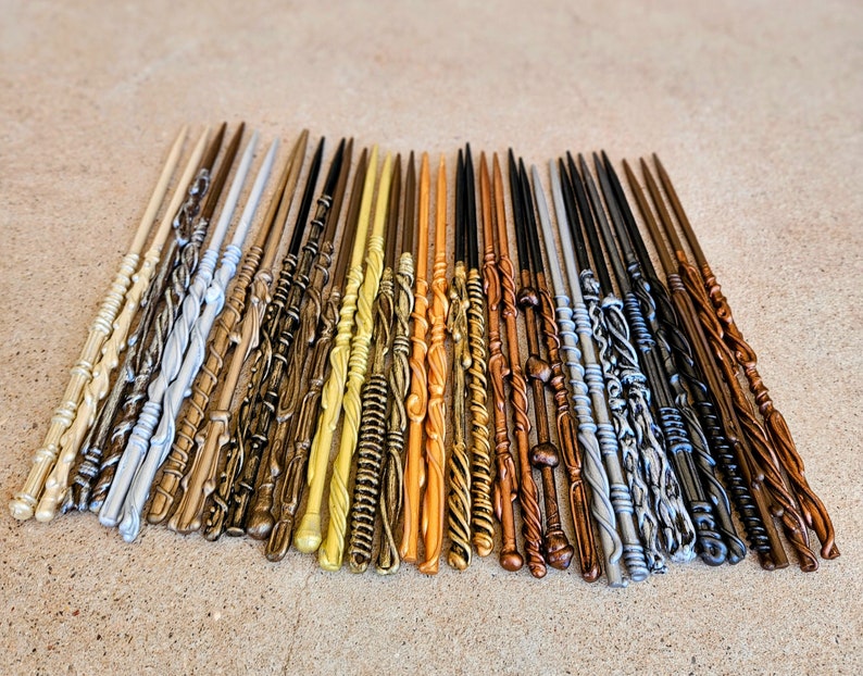 May include: A collection of 25 wands, each with a unique design and color. The wands are made of wood and painted in various shades of gold, silver, brown, and black. The wands are arranged in a row on a light brown surface.