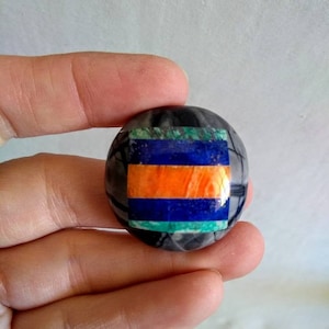 May include: A round, gray stone cabochon with a multi-colored inlay. The inlay is a geometric pattern of orange, blue, and green stones.