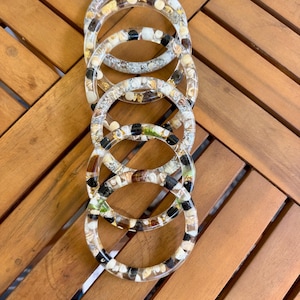 Może przedstawiać: Sześć przezroczystych bransolet z żywicy wypełnionych muszlami, małymi kamieniami i złotymi płatkami. Bransoletki ułożone są pionowo na drewnianej powierzchni, prezentując różnorodność kolorów i faktur. Każda bransoletka ma średnicę około 6,35 cm.