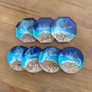 May include: Set of six resin coasters with a beach scene. The coasters are round and octagonal, with blue, white, and brown colors. The coasters feature a beach scene with white sand, blue water, and white shells.