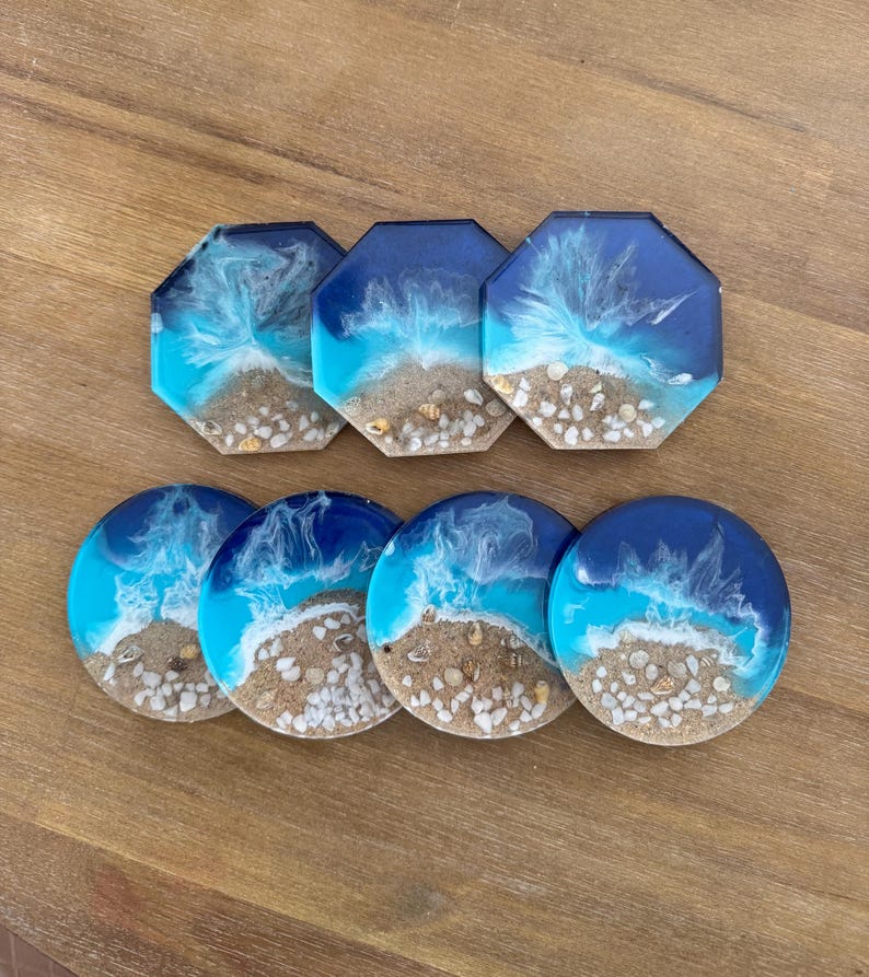 May include: Set of six resin coasters with a beach scene. The coasters are round and octagonal, with blue, white, and brown colors. The coasters feature a beach scene with white sand, blue water, and white shells.