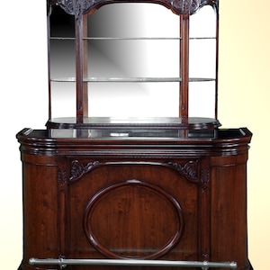 May include: A dark wood bar cabinet with a glass-fronted top shelf and a lower cabinet with a single door. The cabinet has ornate carvings and a silver footrest.