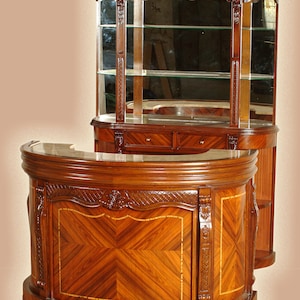May include: A wooden bar cabinet with a curved counter top and a glass-front cabinet with shelves. The cabinet is made of dark wood with intricate carvings and a marquetry design.