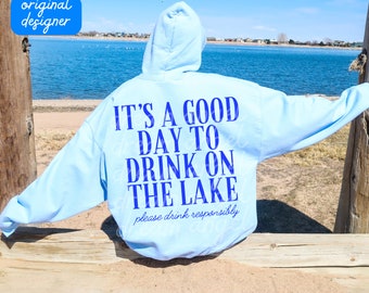 May include: A light blue hoodie with the text "IT'S A GOOD DAY TO DRINK ON THE LAKE" in blue lettering. The text "please drink responsibly" is printed in smaller blue lettering below the main text. The hoodie is worn by a person sitting on a wooden railing overlooking a lake.