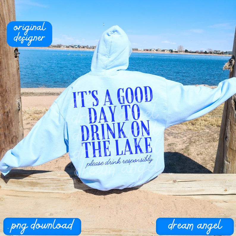 May include: A light blue hoodie with the text "IT'S A GOOD DAY TO DRINK ON THE LAKE" in blue lettering. The text "please drink responsibly" is printed in smaller blue lettering below the main text. The hoodie is worn by a person sitting on a wooden railing overlooking a lake.