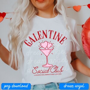 May include: White t-shirt with red text that says "Galentine Social Club" with two pink hearts above a martini glass. The text below the martini glass says "Ladies Celebrating Ladies".