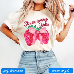 May include: A white t-shirt with the text "Strawberry Girly" in pink and green lettering. The shirt features a watercolor illustration of two strawberries with a pink bow.