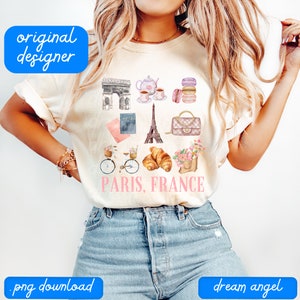 May include: A white t-shirt with a pink and blue design featuring Parisian landmarks, including the Eiffel Tower, Arc de Triomphe, a bicycle, croissants, and macarons. The text "PARIS, FRANCE" is printed in pink on the shirt.