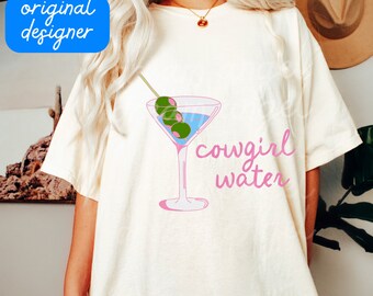 May include: A cream-coloured t-shirt with a pink martini glass design, olives, and the words "cowgirl water" in pink script. The shirt has a relaxed fit and short sleeves. The image also includes the text "original designer", ".png download", and "dream angel".
