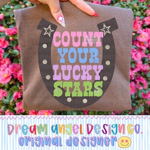 Peut inclure: Un t-shirt marron avec un motif de fer à cheval et le texte "COUNT YOUR LUCKY STARS" en rose, bleu, violet et vert. Le design comprend des étoiles et des paillettes. Le bas du t-shirt porte l'inscription "Dream Angel Design Co. Original Designer".
