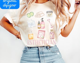 May include: A white t-shirt with the word "ESTHETICIAN" printed in pink and a watercolor illustration of various spa and beauty products, including a face mask, cucumber slices, a bottle of lotion, and a jar of face cream.