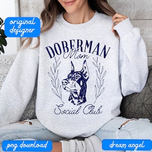 Puede incluir: Una sudadera gris con un diseño gráfico azul que dice "Doberman Mom Social Club". El gráfico presenta la cabeza de un perro Doberman Pinscher con un esquema de color azul y blanco.