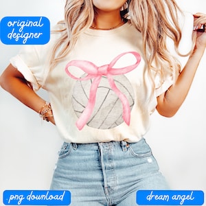 Può includere: Una maglietta bianca con un'illustrazione ad acquerello di una pallavolo con un fiocco rosa in cima.