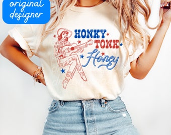 May include: Cream-colored t-shirt featuring a vintage-style illustration of a cowgirl playing a guitar, with the words "Honky Tonk Honey" in red and blue. The design includes stars and a rope-style font.