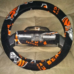 May include: Black steering wheel cover with orange and white Cincinnati Bengals team logos and text. The cover features a repeating pattern of helmets and the team name. The cover is designed to fit a standard steering wheel.