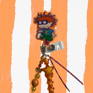 Könnte beinhalten: Ein einziehbarer Ausweis-Clip mit einer Zeichentrickfigur mit orangefarbenem Haar und Brille, die ein blaues Hemd und eine grüne Hose trägt. Die Rolle hat einen Perlenanhänger mit orangefarbenen und gelben Perlen sowie lila und orangefarbenen Stäben. Der Hintergrund ist orange und weiß gestreift.